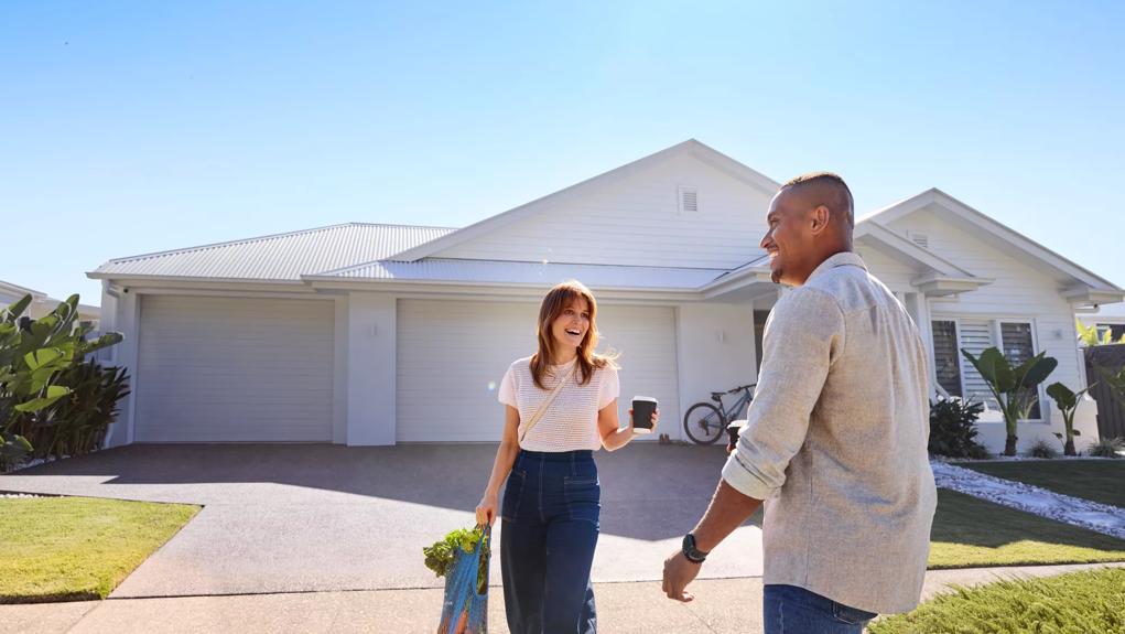 Young couple standing outside their home on the driveway, smiling, groceries in hand.