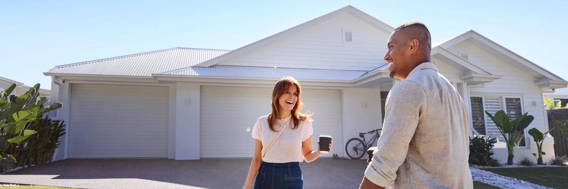 Young couple standing outside their home on the driveway, smiling, groceries in hand.