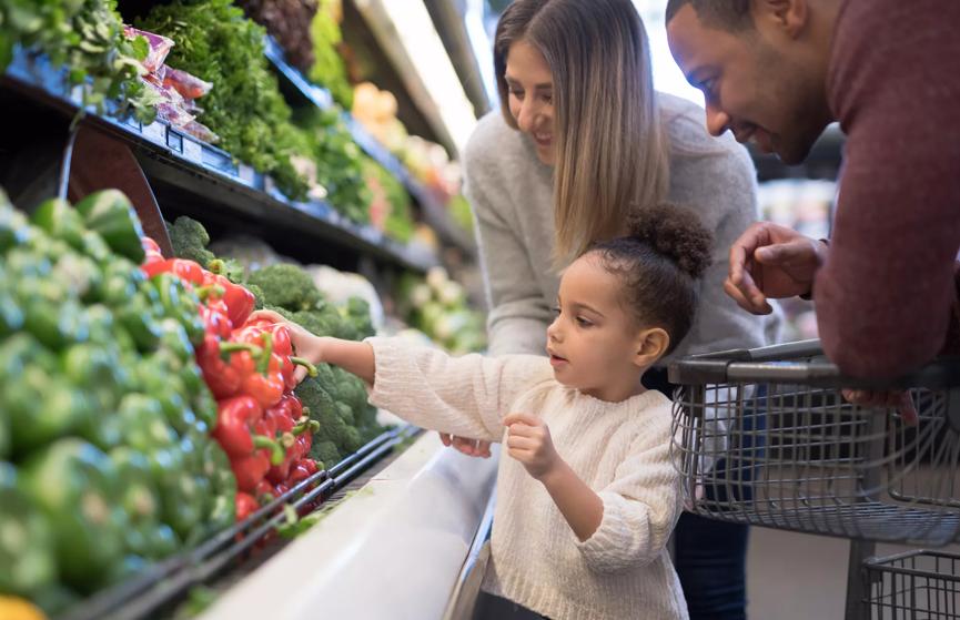 A pre-school age girl helps her parents pick out veggies in the produce section at the grocery store. She is reaching for a red bell pepper. Her mother is Caucasian and her father is African-American.