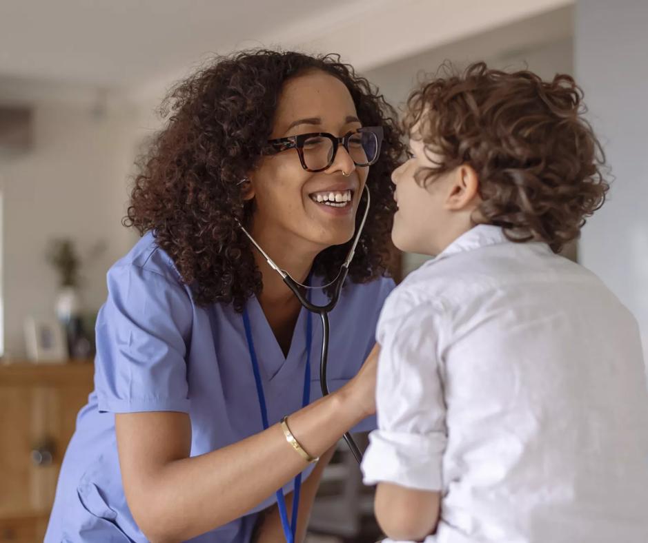 A female doctor smiles while checking the heart and lungs of a pediatric patient during a medical appointment.